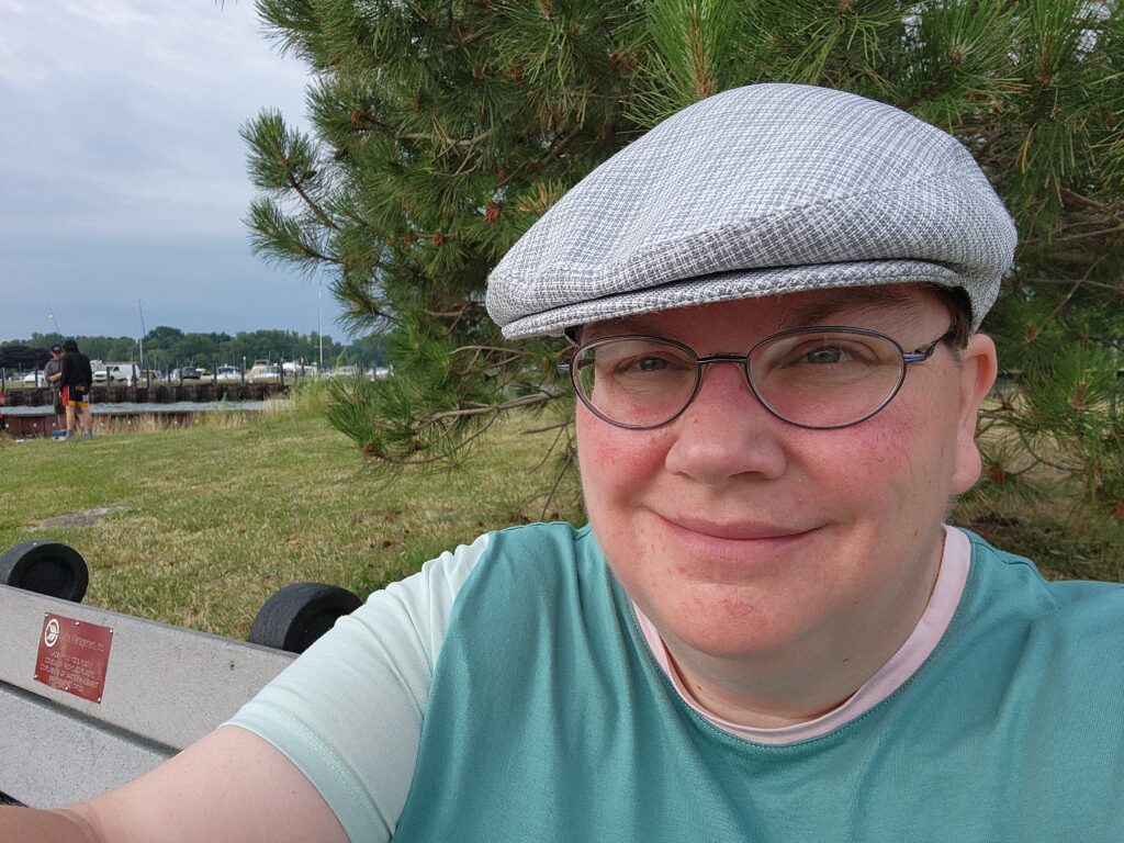 Selfie of me sitting on a bench at the boat launch at 9-Mile in St Clair Shores. There's an evergreen tree behind me and a couple of young anglers and some boats on a nearby dock behind me.